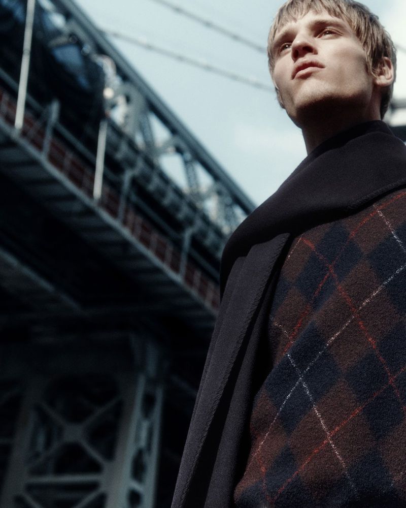 Portrait of a blond male fashion model outdoors with a modern urban bridge, wearing a plaid sweater and dark coat, looking up.