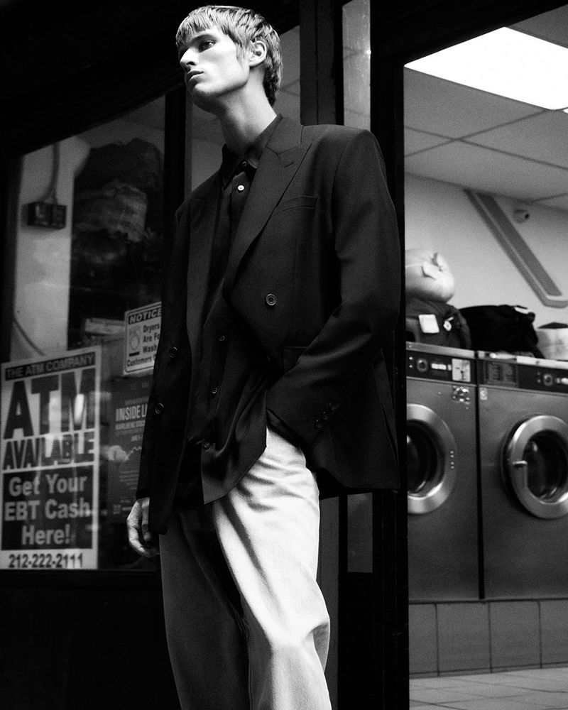 Black and white shot of a man in a suit standing outside near a laundromat, creating an editorial fashion portrait.