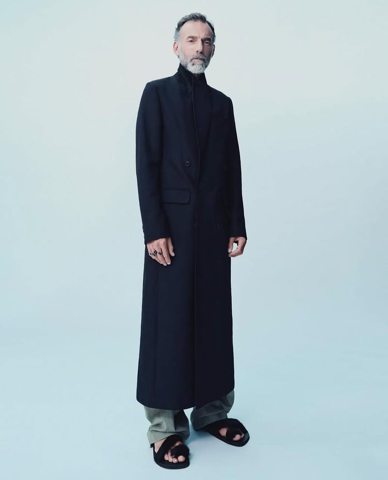 A man in a black coat, light-green pants, and black sandals stands in a brightly-lit white studio, looking directly at the camera.