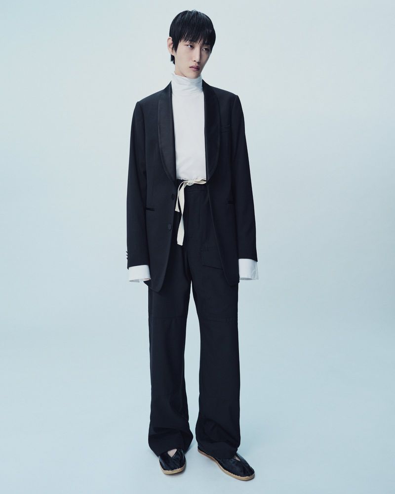 A solo Asian male fashion model poses in a black suit with a white turtleneck indoors against a light background in a full shot.