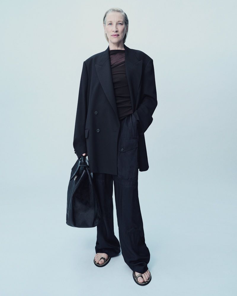 Older woman stands in black blazer, top, and pants, holding a large black bag in a studio setting with a light blue background.