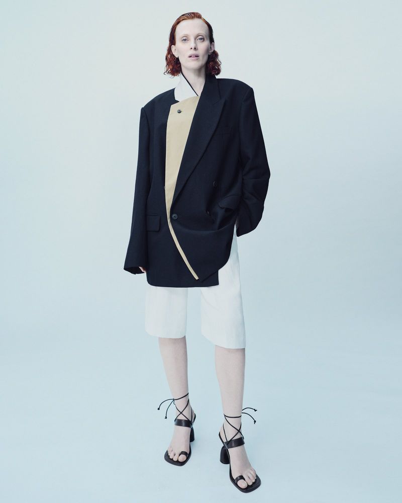 Adult woman in an oversized black blazer, white shorts, and wrap-around heeled sandals poses in a brightly lit photo studio setting.