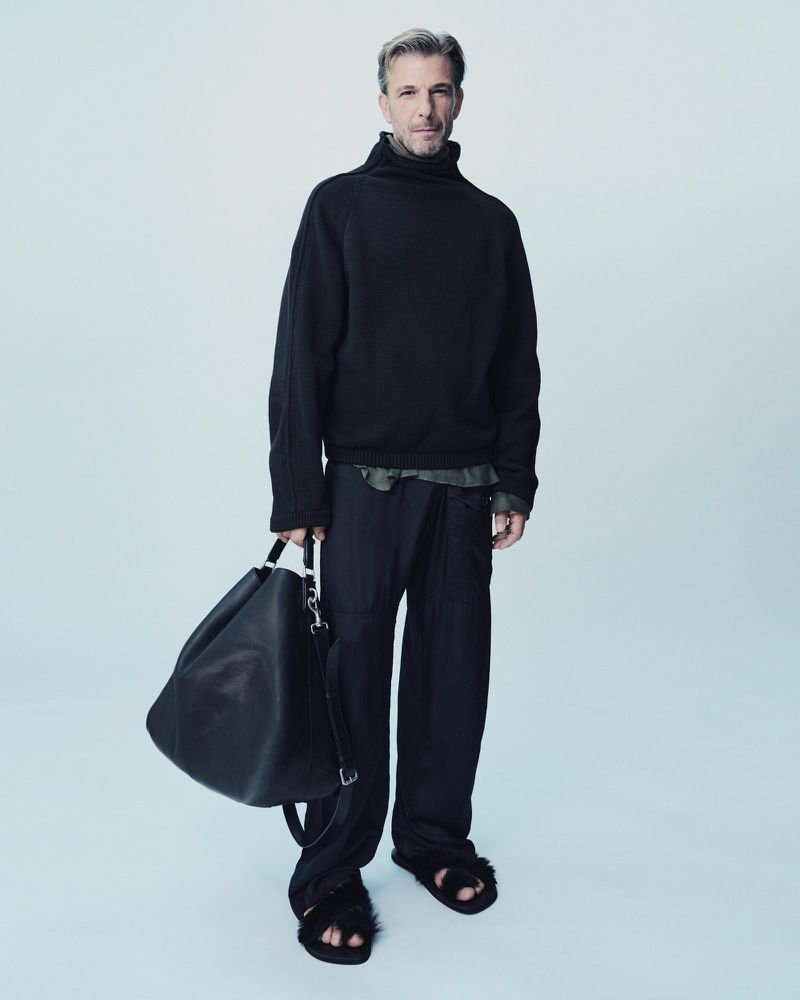 Full-length studio shot of a handsome man in a black turtleneck sweater, loose black pants, and sandals holding a black bag.