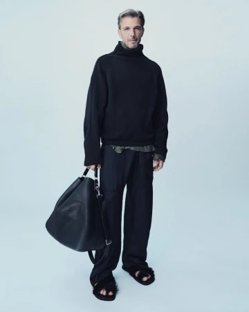 Full-length studio shot of a handsome man in a black turtleneck sweater, loose black pants, and sandals holding a black bag.