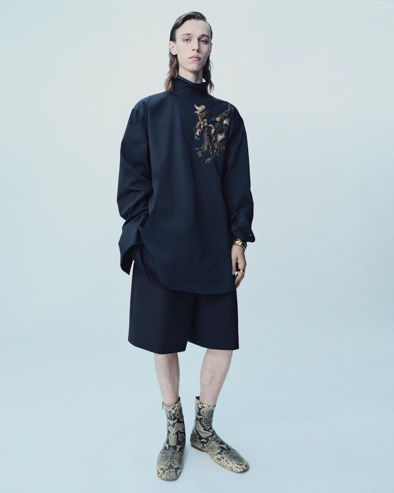 Model posing in an oversized black high-neck tunic with gold embroidery, black shorts, and printed boots against a pale seamless background.