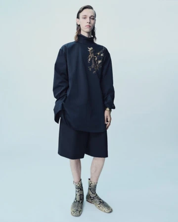 Model posing in an oversized black high-neck tunic with gold embroidery, black shorts, and printed boots against a pale seamless background.
