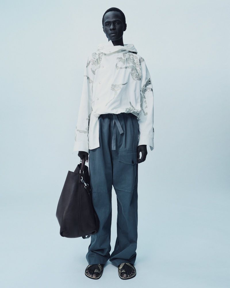 Full-length shot of a dark-skinned adult male wearing a white hooded shirt and blue trousers, holding a dark-brown bag, studio lit.