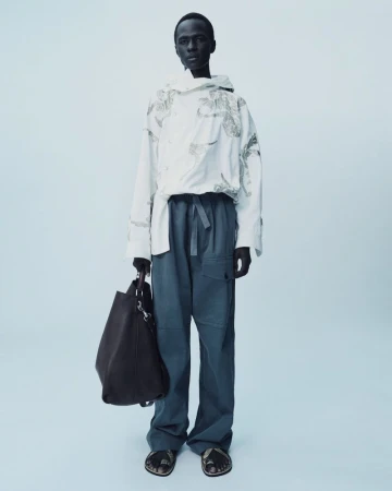 Full-length shot of a dark-skinned adult male wearing a white hooded shirt and blue trousers, holding a dark-brown bag, studio lit.
