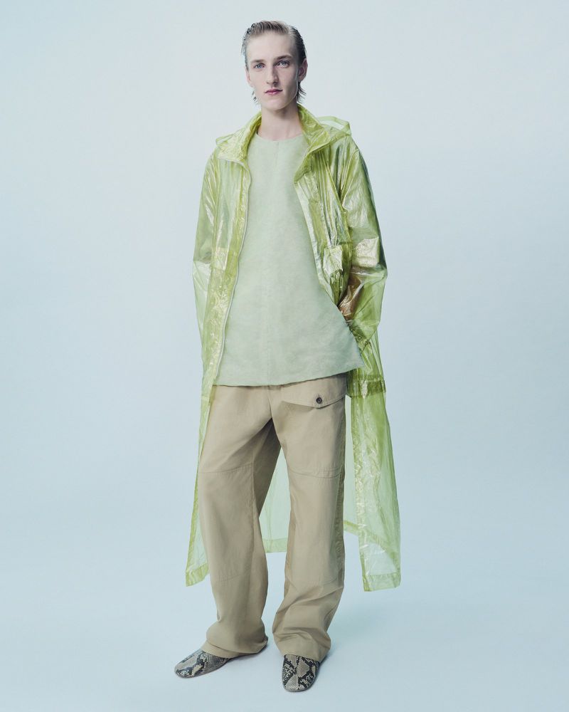 Full-shot of a slender adult man in a shiny yellow hooded raincoat, beige pants, and snakeskin-print flats, posing in a photo studio.