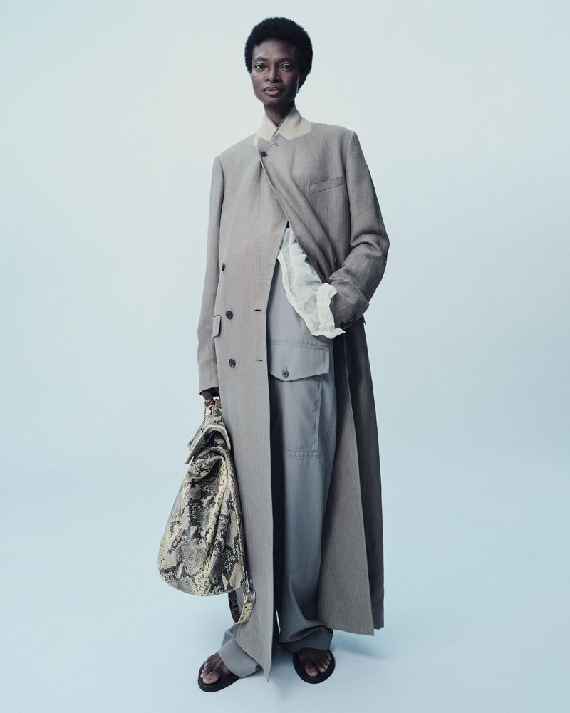 A full-length fashion portrait of an adult female model in a neutral overcoat, grey pants, and patterned bag against a studio background.