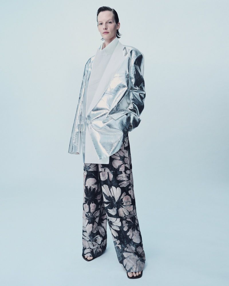 Fashion shot of a woman in silver jacket and floral print pants standing against a light blue backdrop in a photo studio setting.