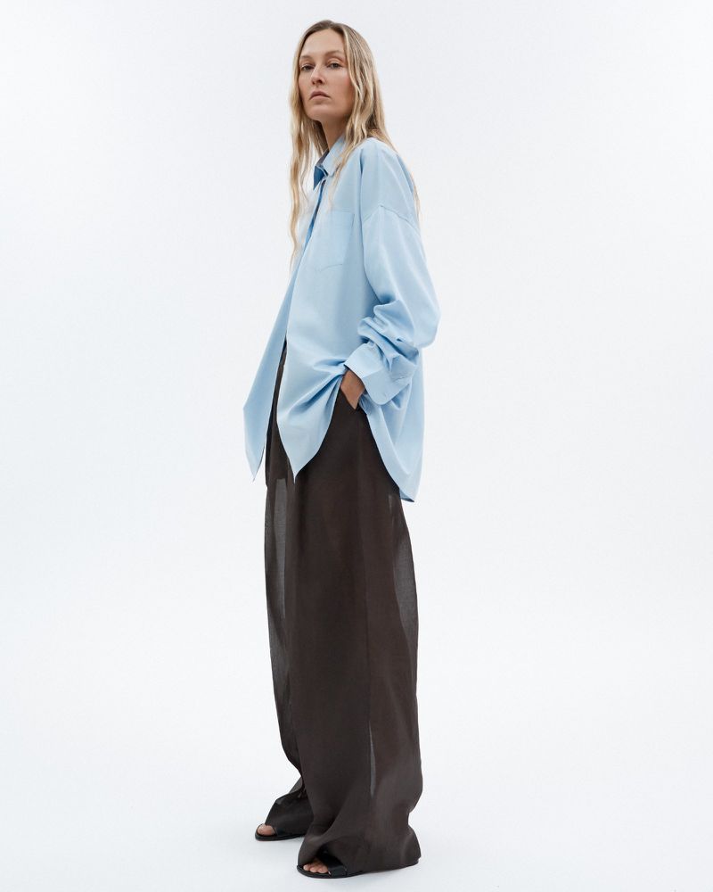A blonde woman poses in a light blue button-down shirt and dark, loose-fitting pants against a clean, white backdrop in a fashion shot.