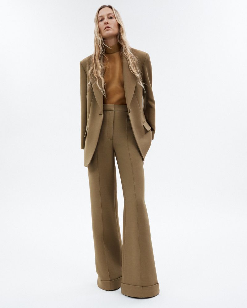 Adult woman in a neutral monochrome fashion look with a blazer, sweater and wide leg trousers in a photo studio against a white backdrop.