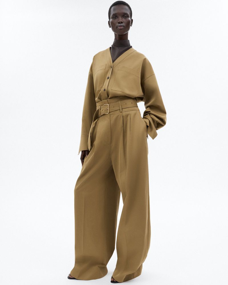 Elegant dark-skinned woman in a tan blouse and wide-leg belted trousers posing against a bright white backdrop in a fashion editorial.