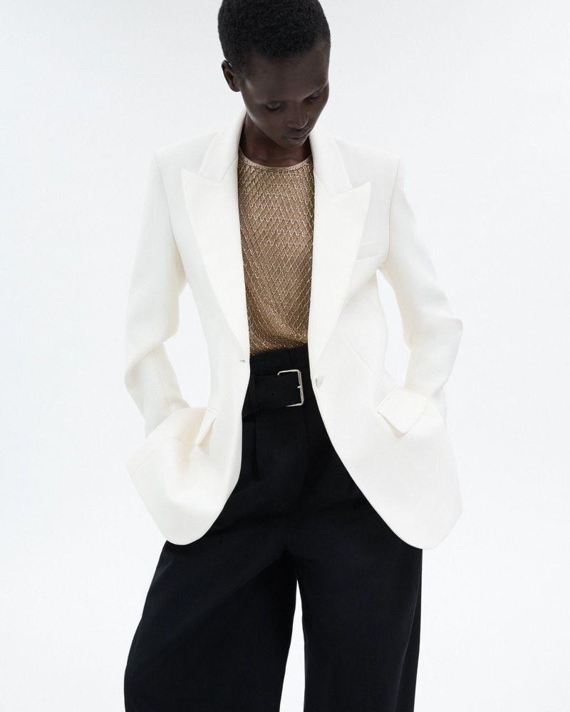 Fashionable woman in a white blazer, gold top, and black pants stands confidently against a bright backdrop in a studio shot.