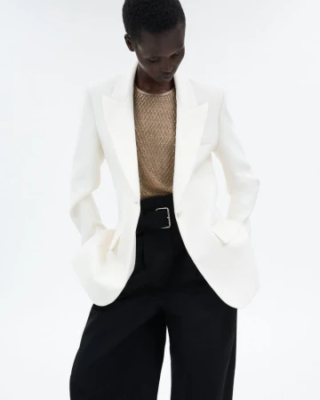 Fashionable woman in a white blazer, gold top, and black pants stands confidently against a bright backdrop in a studio shot.