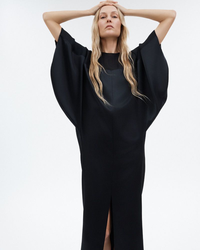 Elegant woman in a full-length black dress poses against a clean white backdrop with hands resting on her head in a fashion-forward image.