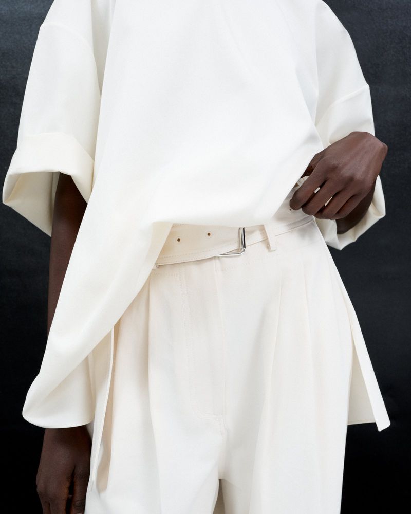Closeup of a light-skinned human wearing a creamy-white oversized top and matching belted pants against a dark grey background.