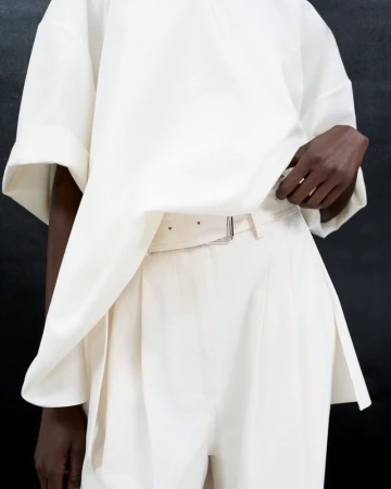 Closeup of a light-skinned human wearing a creamy-white oversized top and matching belted pants against a dark grey background.