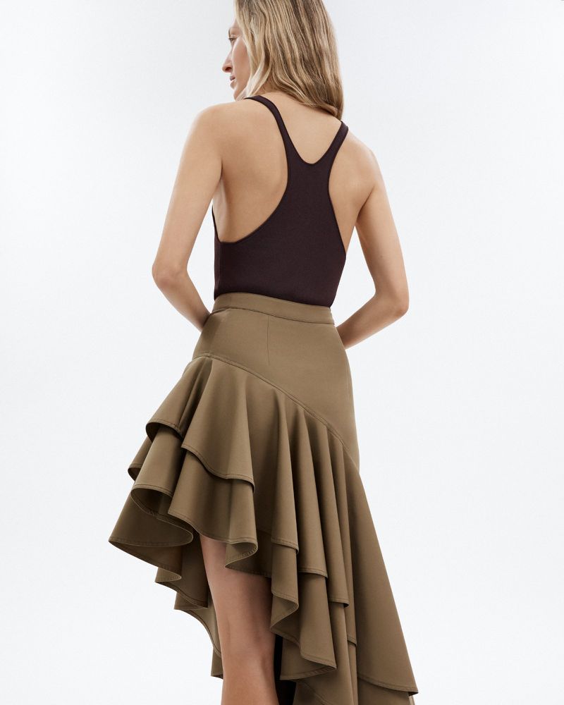 Blond adult woman wears a brown bodysuit and a tiered olive green asymmetrical skirt. The background is bright, even, and light.