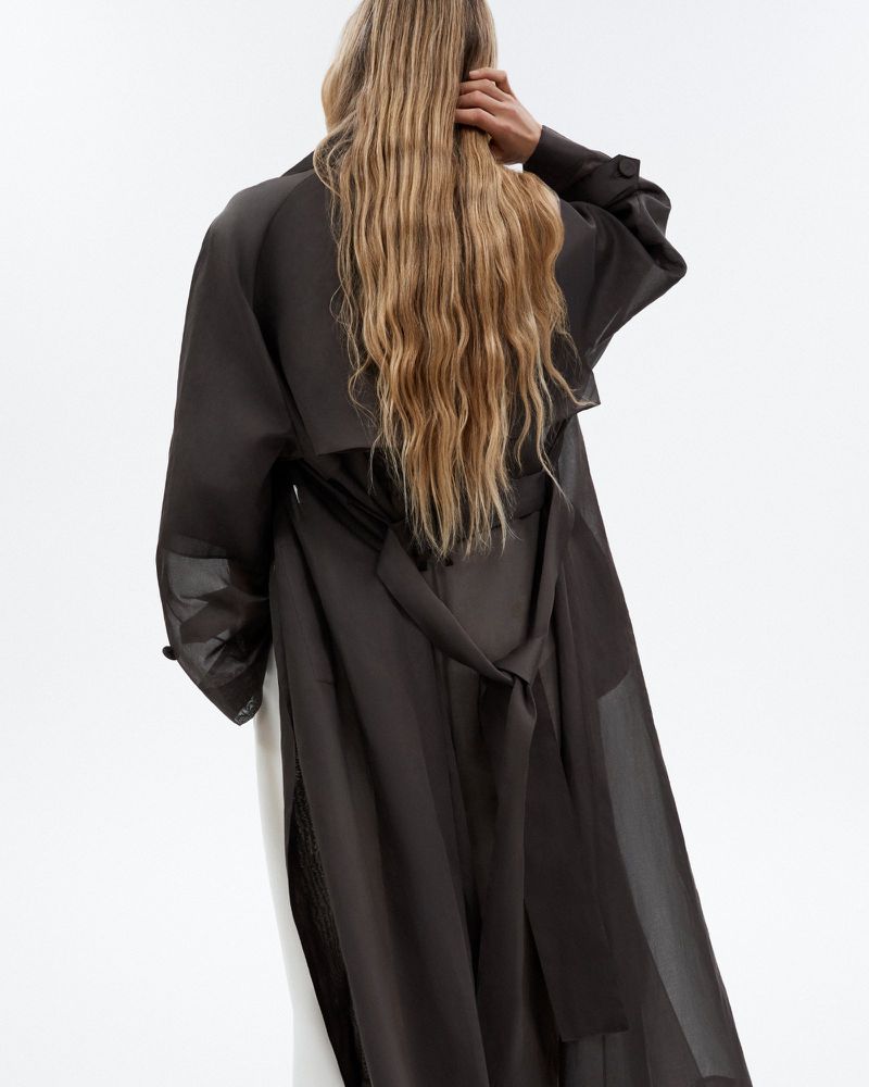 Back view of an adult female with long blonde hair in a long dark grey trench coat with white pants in a studio setting.
