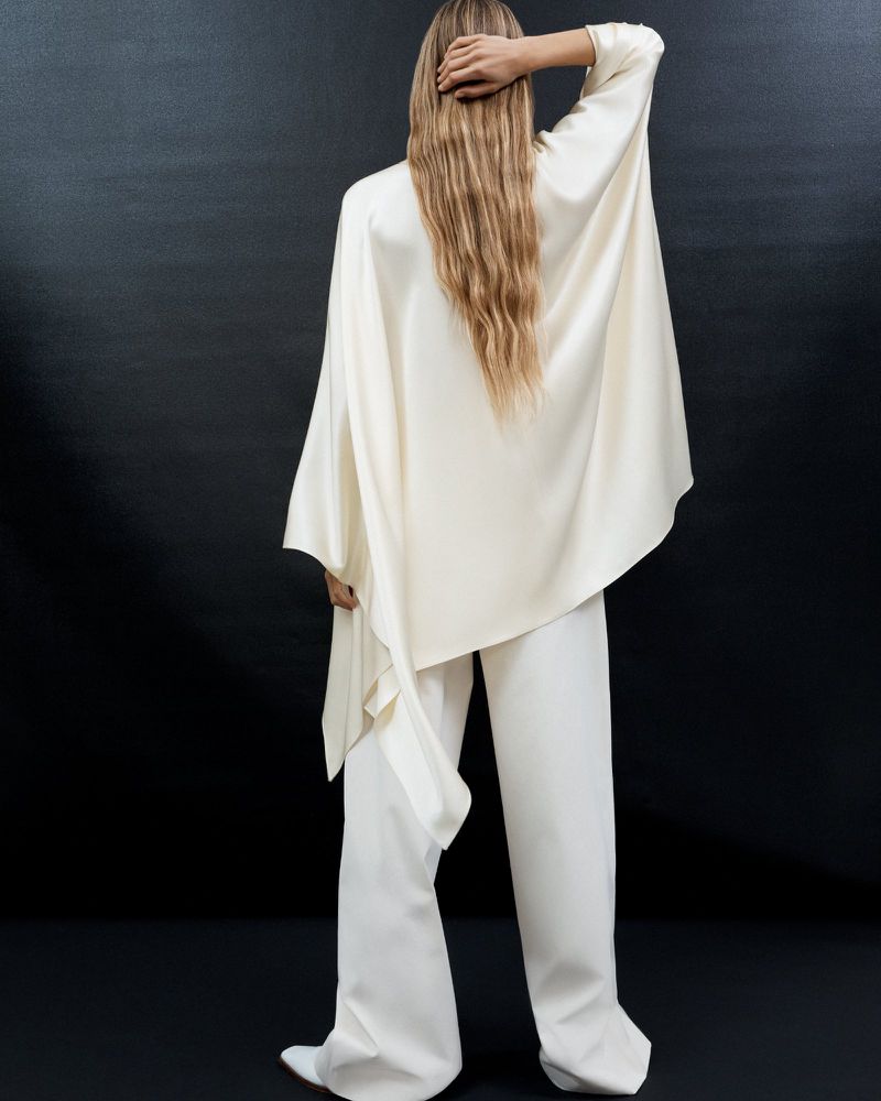 Back view of a tall woman wearing an off-white cape-sleeved top and wide-legged pants in a studio setting with a dark background.