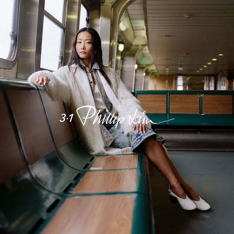 An Asian woman in an off-white coat is sitting on a bus seat, posing for a fashion editorial shot with blurred background, medium shot.