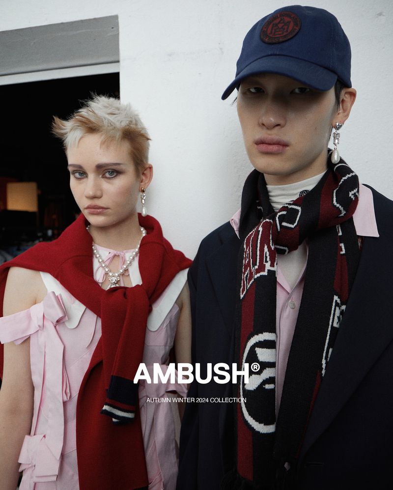 Two fashion models wearing apparel from the Ambush Autumn Winter 2024 collection, standing in front of a plain wall backdrop.