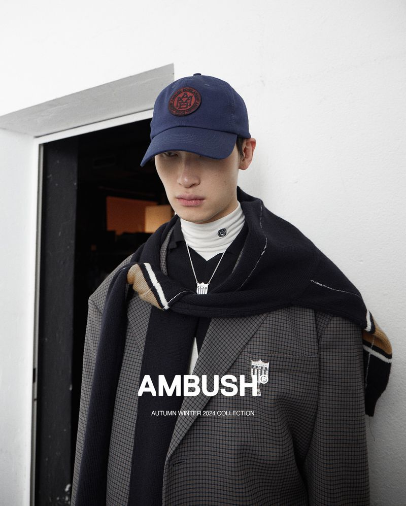 Stylish young man in a cap, blazer, and scarf from the Ambush Autumn/Winter 2024 collection, posing in an indoor setting.