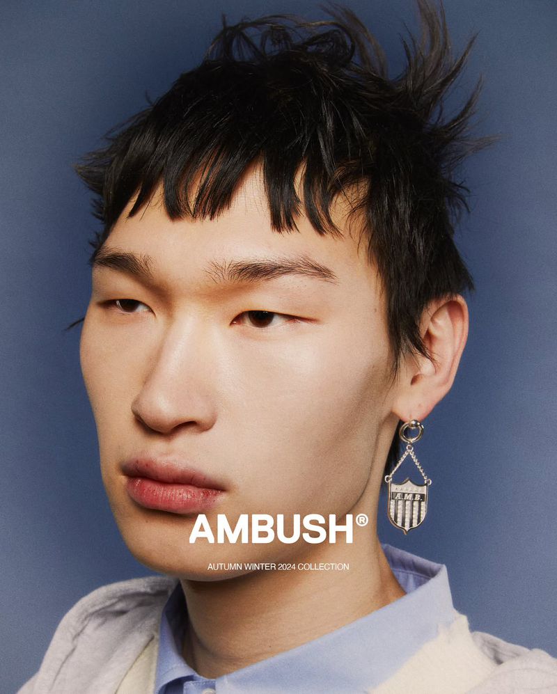 Close-up studio portrait of a male model wearing an earring, showcasing fashion, set against a blue backdrop with the Ambush brand logo.