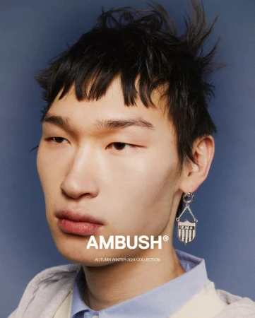 Close-up studio portrait of a male model wearing an earring, showcasing fashion, set against a blue backdrop with the Ambush brand logo.