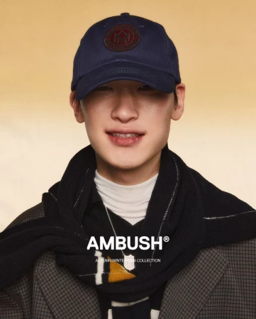 Portrait of Asian male model in a baseball cap and black scarf, showcasing the Ambush Autumn Winter Collection against a warm backdrop.