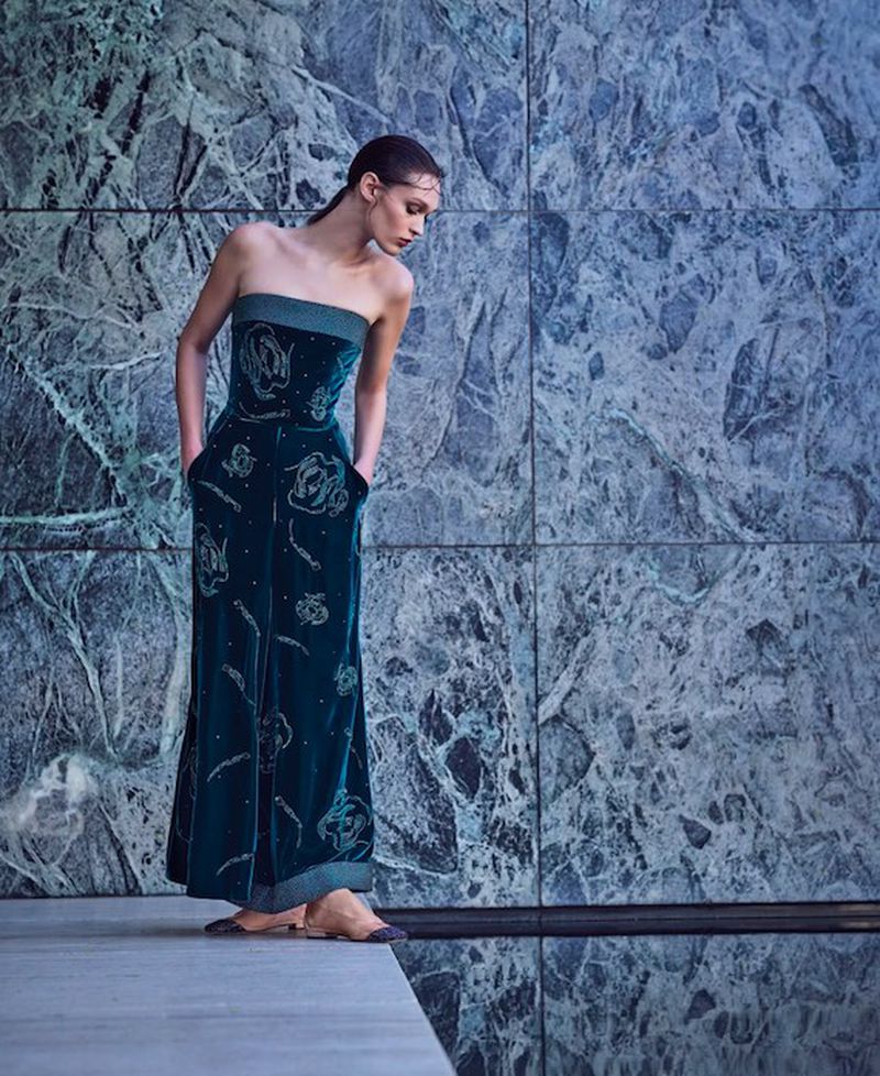 A woman poses in an elegant teal jumpsuit with floral embellishments against a textured, marbled wall with reflective flooring.