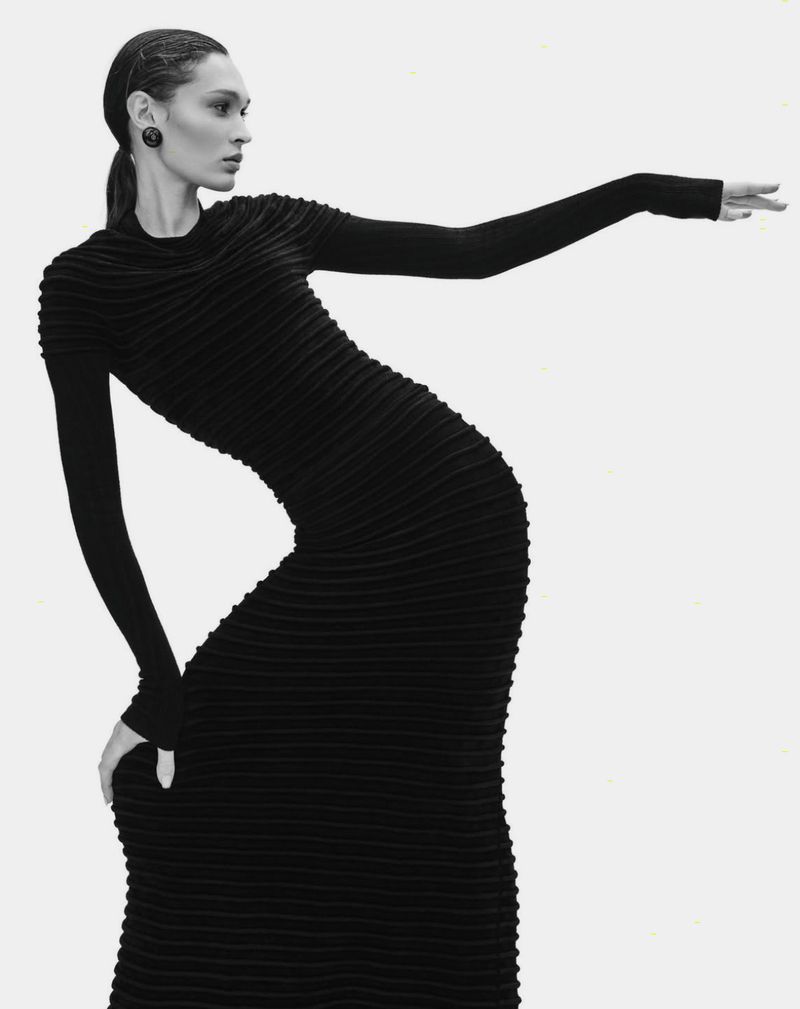 Fashion shot of a slender woman in a black textured gown, posing with outstretched arm against a stark white background in studio lighting.