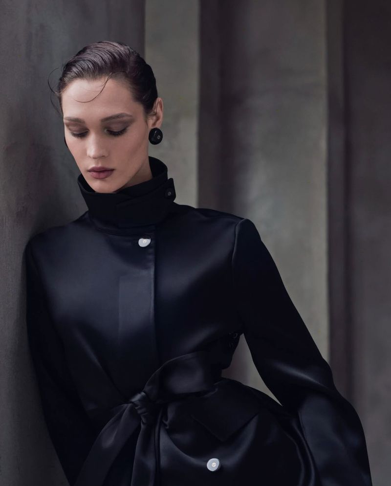 Elegant portrait of a brunette woman in a black high-necked satin coat with button details, leaning against a gray architectural wall.