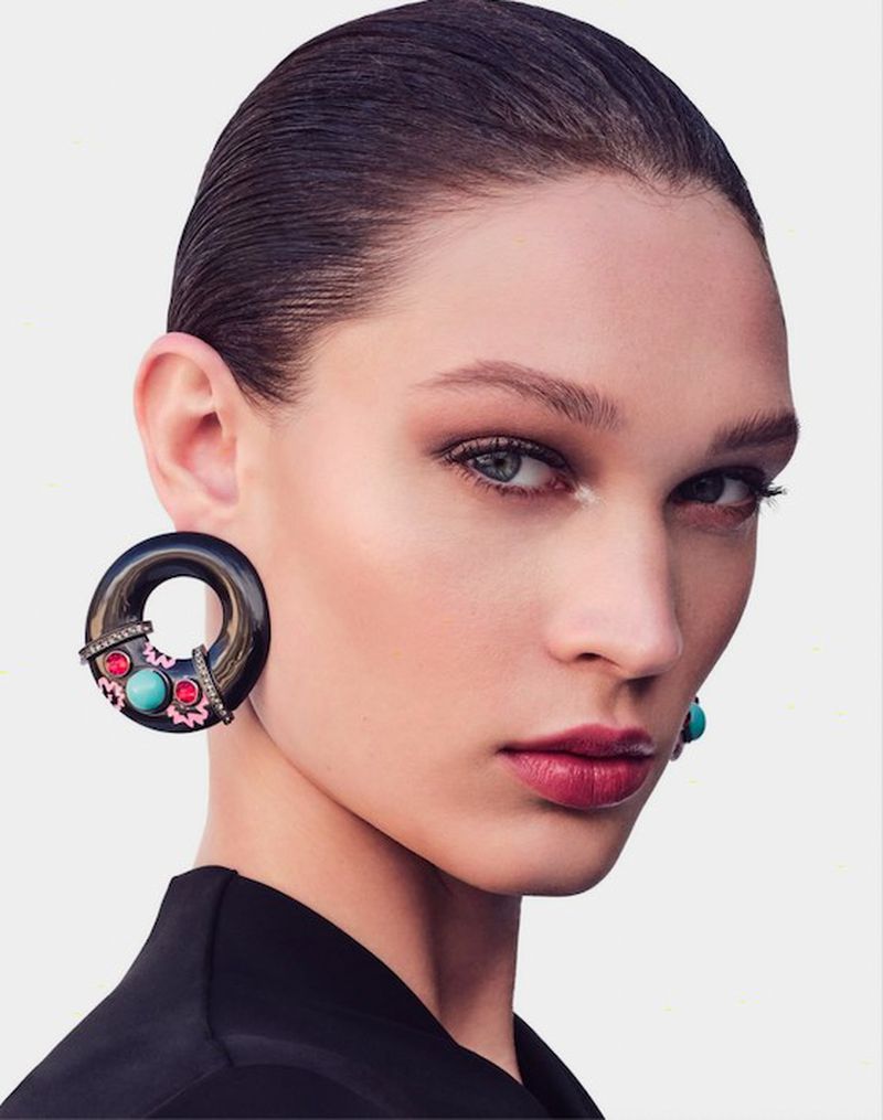 Close-up of a woman with dark hair, wearing an ornate black, turquoise, and pink earring against a plain white backdrop.