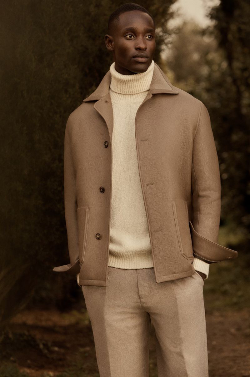 A dark-skinned male model poses in an outdoor setting wearing a tan jacket, cream turtleneck sweater, and tan dress pants for a fashion editorial.