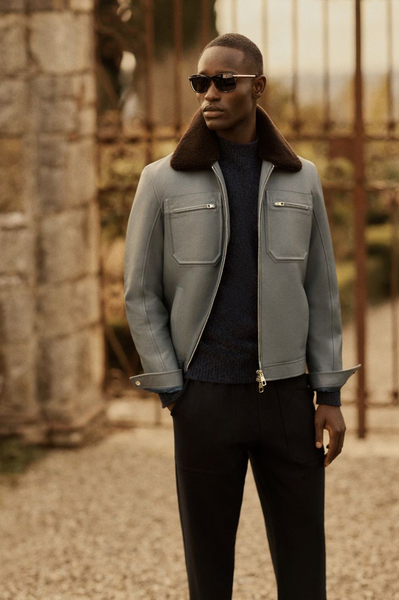 Charles Oduro wears a grey zip jacket with a brown shearling collar and dark sunglasses in front of an iron estate gate.