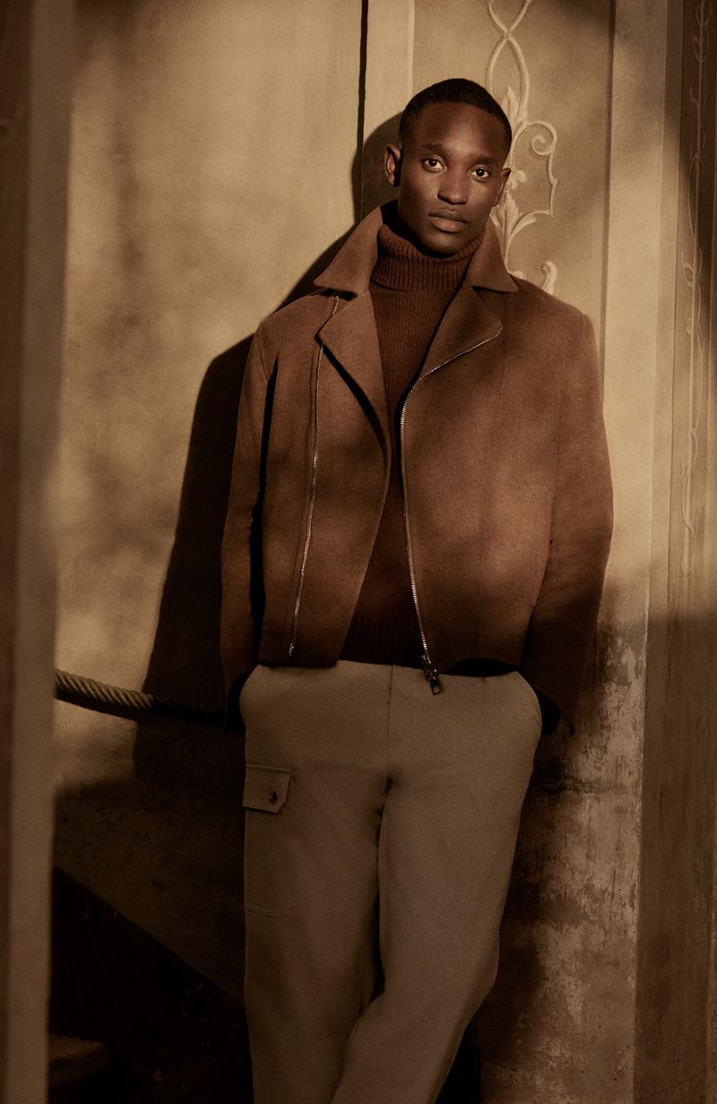 Fashionable man in a brown jacket and tan pants stands in a sepia-toned indoor setting with architectural details, posing for a portrait.