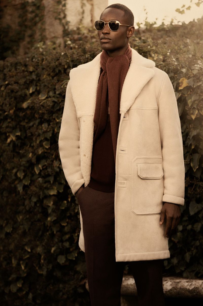 A fashionable Black man in a cream-colored coat and burgundy scarf stands outdoors against a leafy green background, gazing off-camera.