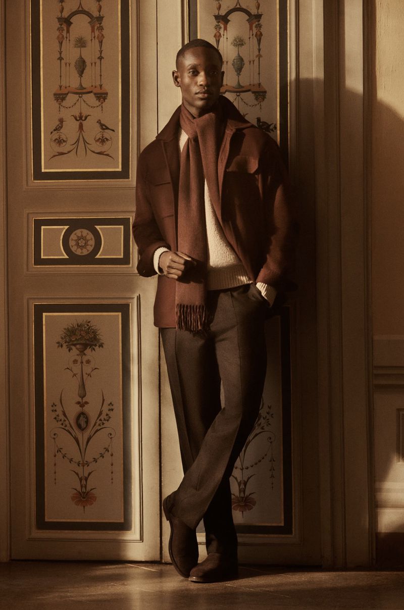 Portrait of a dark-skinned adult man wearing a brown jacket and scarf, leaning against an ornate door inside. Warm, inviting fashion shot.