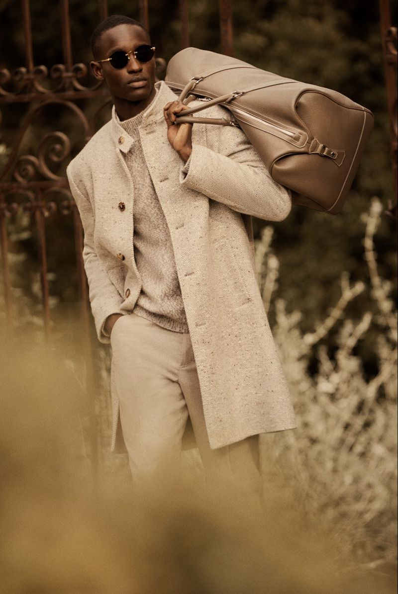 Man in a beige coat and round sunglasses carrying a large duffel bag stands outdoors against a blurred leafy background and wrought-iron gate.
