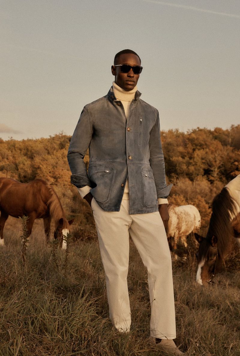 A stylish Black male model poses in a denim jacket and neutral pants in a rural landscape with horses, bathed in warm, natural light.