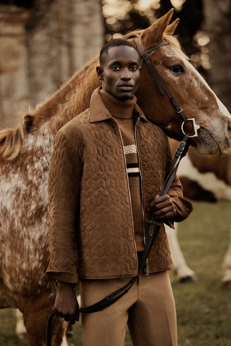 A stylish African male model in a quilted jacket poses with a chestnut horse outdoors, exuding sophistication and rural elegance.