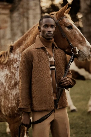 A stylish African male model in a quilted jacket poses with a chestnut horse outdoors, exuding sophistication and rural elegance.
