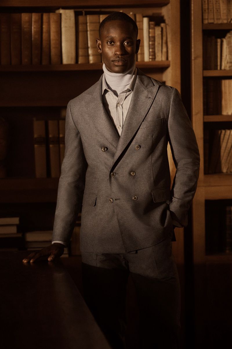 Portrait of an adult male in a gray suit, posing in a library with wood bookshelves filled with books, casting a strong shadow.