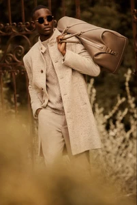 Black man wearing sunglasses and light tweed coat holding leather duffel bag outdoors for Brett Johnson lookbook