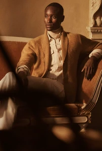 Model seated on a classic sofa wearing a tan cardigan sweater and white trousers for the Brett Johnson Fall/Winter 25 lookbook.
