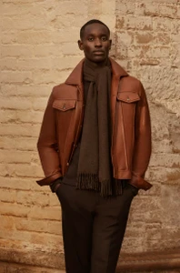 Model Charles Oduro wearing a brown leather jacket and dark wool scarf standing against a rustic brick wall for Brett Johnson FW25.
