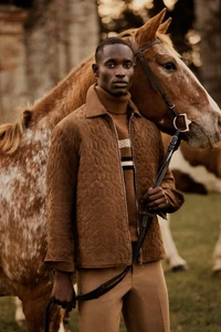 Model Charles Oduro wears a brown quilted jacket and camel trousers while holding a brown horse on a grassy field.